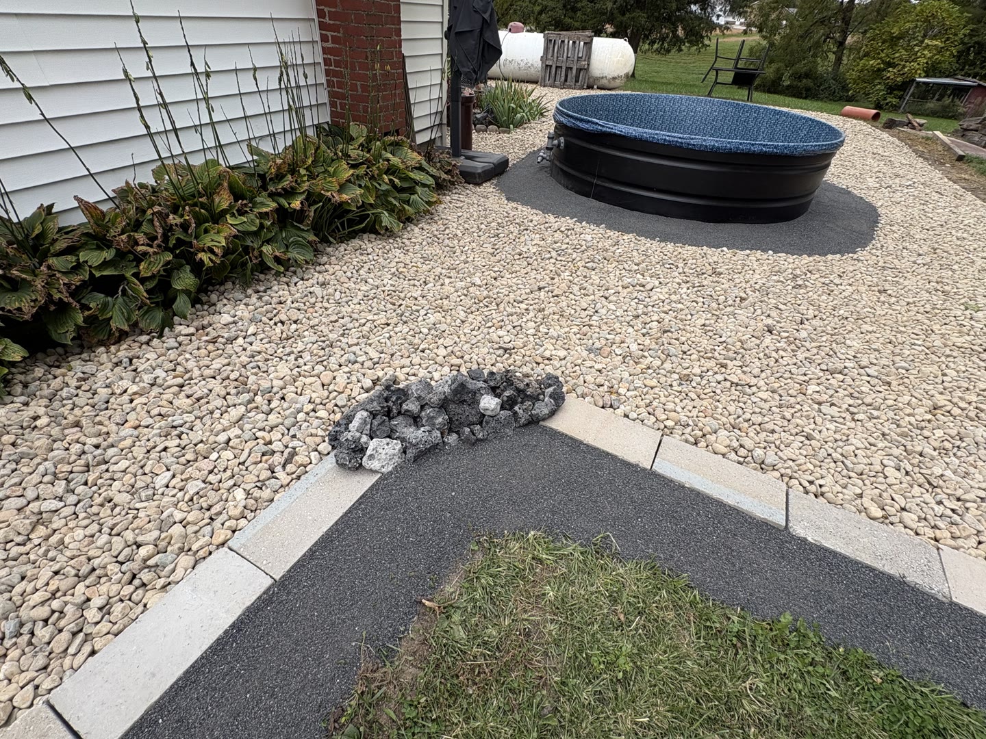 A real garden with a dark mineral border and rough stone accents beside grass and a round garden tank; separate light rounded gravel fills the adjacent bed.